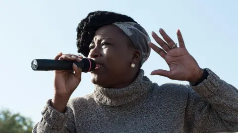 Shola Mos-Shogbamimu Shola Mos-Shogbamimu photographed outside, against a blue sky, with a hint of a tree in the background in the bottom left hand corner. She is looking off-camera, towards the left of the frame and is holding a microphone in her right hand. She is pictured giving a speech and is mid-sentence. Her other hand is emphasising what is being said - held up to her head height, with her fingers spread and palm showing. She is wearing a rollneck black, white and grey sweater.
