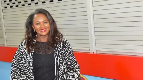 Emertha Uwanyingira is a middle aged Black woman of Rwandan heritage. She has long brown hair and is wearing a black top with a loose black and white print jacket over it. One earring is visible in her right ear. She is wearing red lipstick and smiling at the camera. She is standing in front of a red counter with silver shutters pulled down over it. It looks like a canteen.