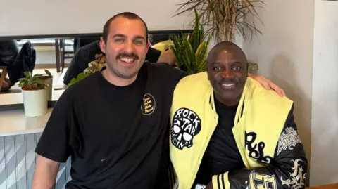 Aidan Jackson wearing a black t-shirt poses with Akon who is wearing a yellow, black and gold jacket. The jacket has a logo which reads "Rock Star Original". 