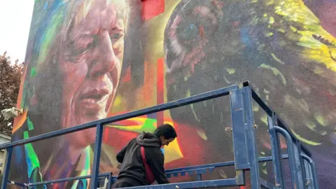 Upfest HazardOne is designing Sir David Attenborough's mural. She is standing on a blue platform. HazardOne has black hair and is wearing a grey jumper and red body bag. 