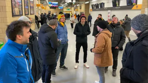 Dean Sullivan The friends stand on the concourse inside the Wolves ground. They have coats and jackets on and some have bobble hats on and are smiling together.