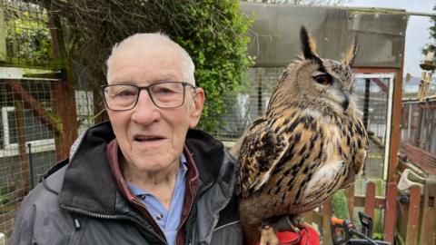 An elderly man with white hair and glasses holding an owl beside his head