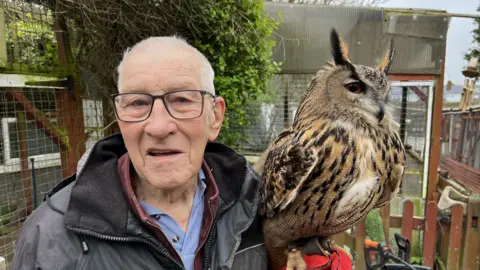 BBC An elderly man with white hair and glasses holding an owl beside his head