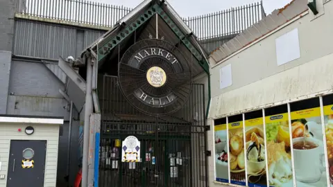 The entrance to Barrow Market Hall which has a black fence in front of it. The roof is pointed and there is a large circular sign towards the top with a coat of arms and old lettering which reads 'Market Hall'. There is a sign stuck on the gate which has black and yellow tape surrounding it. There is a Herons shop to the right which multiple pictures of food on the side of the wall.