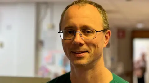 BBC/Gill Dummigan Dr Michael Stewart, wearing green scrubs, stands in a hospital corridor