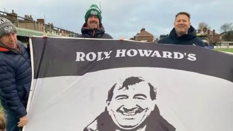 Three men hold a large black and white flag with an image of Roly Howard. Out of shot are the words: "Roly Howard's Black and White Army".