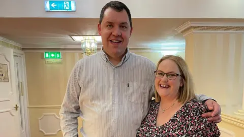 Lorraine Wallace Two people stand together in a hotel or function venue corridor with cream striped walls and a red patterned carpet. One person wears a light striped button-down shirt and jeans, while the other wears a dark floral dress and glasses.