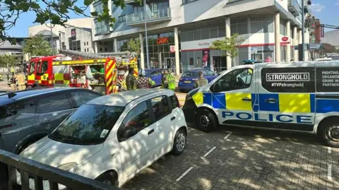 A fire engine and police van parked outside a post office. There is another white car and black car parked next to them. There are emergency workers standing outside.