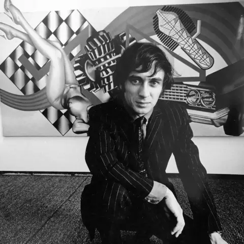 Courtesy of the Phillips family A black and white image of Peter Phillips in front of one of his distinctive artworks. He is wearing a dark pinstriped suit, tie and shirt. He is crouching down in front of the painting. 