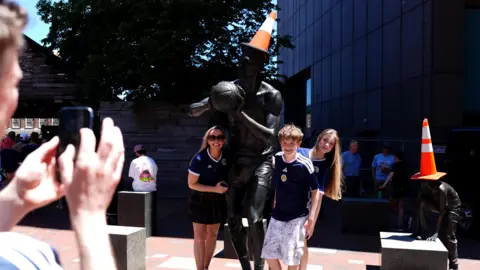 PA Media A man taking a photograph of two women and a boy wearing blue Scotland football shirts standing next to statues with orange traffic cones on their heads.
