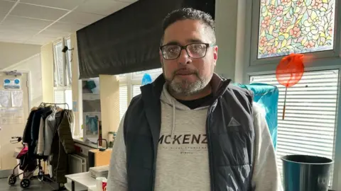 BBC A man wearing a grey jumper and a navy gilet stood in a community centre. he wears black-rimmed glasses and has tight curled black hair. Behind him are windows with blinds on them, a coat rail and a white table.