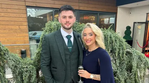 Charlotte Gallen Man and a woman stand side by side. The woman, on the right, wears a navy dress with a gold necklace. She has blonde hair and make up on. She holds a glass of champagne in her left hand which has a gold watch on it. The man has a green tweed suit with green waistcoat and green tie. He has a white shirt and a tie bar on. He has short brown hair and a short brown beard. Behind them is a wooden building with large glass windows. A green bush sits between the couple and the building