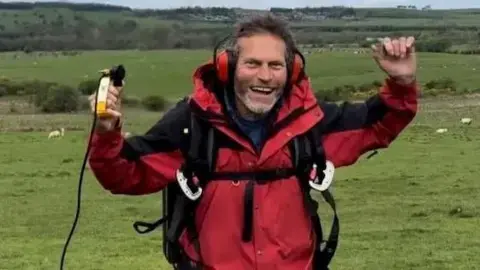 Malcolm Grout, wearing ear protectors, a parachute and a red coat, stands and smiles in a field.