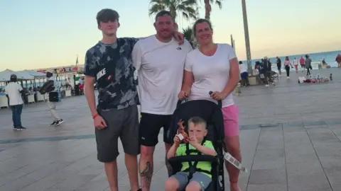 Family handout Alex's family are posing for a picture as the sun sets while on holiday. Alex is standing on the far left with his arm around his father. His mother is standing on the far right and she has her hands on a pram where his brother is sitting holding a toy dinosaur.