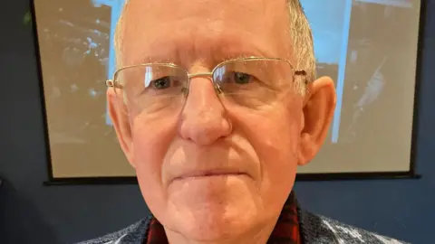 Barry Lycett is wearing glasses and smiling. He is wearing a jumper and a shirt underneath and standing in front of a photo on a wall.