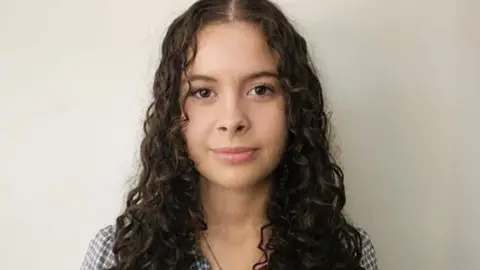 Handout A teenager with long curly brown hair is looking ahead with a slight smile. She is wearing a chequered shirt. 