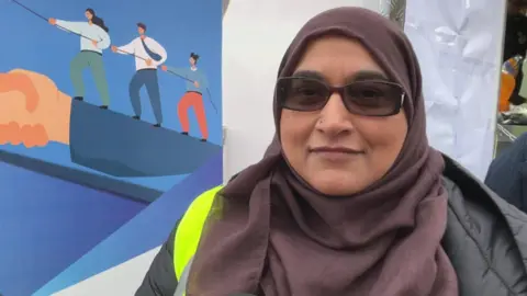 Aisha Iqbal/BBC A woman in a brown headscarf, green coat and wearing a  yellow hi-vis jacket.