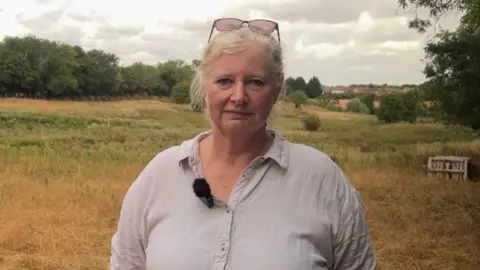 BBC/Lilah Parsons Sue Nodder wearing a light-coloured button-up shirt standing in a nature reserve. Dry grassland, trees, and open countryside extend into the background under a cloudy sky. A clip-on microphone is attached to her shirt, and glasses rest on top of her head.
