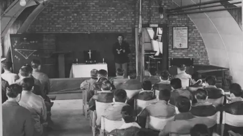 Supplied A black and white photo of rows of men sitting on chairs. In front of them is a table with a cross on it. A man at the front is holding a Bible while addressing the congregation.