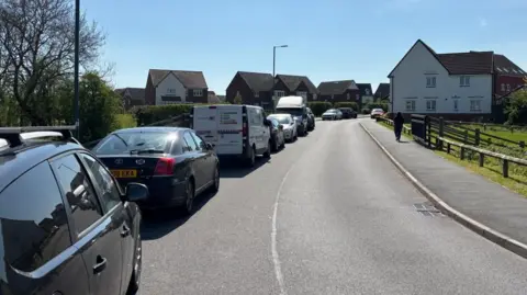 Charlie Stubbs / BBC There is a road with a blind bend either side. To the left, there is a row of cars and work vans. In the foreground, there are dark red bricked and white houses. 