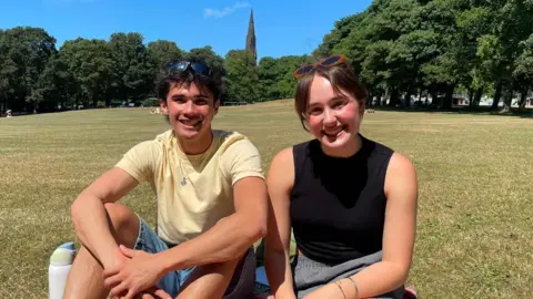 Two people sat in a park on a bright sunny day. On the left, a young man smiles at the camera as he wears a loose fitting yellow t-shirt and denim shorts. To the right, a young woman smiles at the camera in a black vest top. She wears sunglasses in her brunette that she wears tied up.