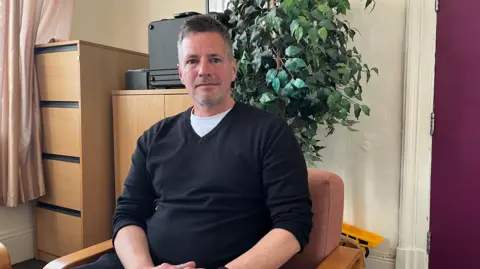Clare Worden/BBC Paul Lambert is sitting in an wooden chair with a green pot plant behind him. He has short black hair. He is wearing a black jumper with a white t-shirt underneath.