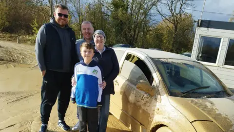 Sally Sarl Sally with her two sons and young grandson standing next to a muddy rally car. Sally is wearing a head scarf, jeans and a navy jumper. Her family are all wearing dark tracksuit bottoms and hoodies. 