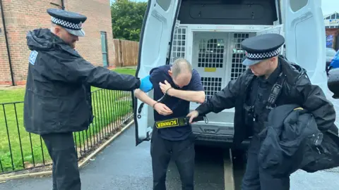 Photograph of a man being scanned for weapons by two police officers. the image shows the suspect before he is bundled into the back of a police van.