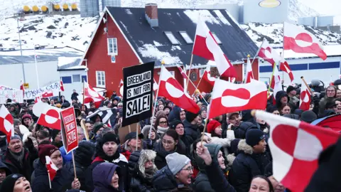 Many protesters in Greenland holding flags and placards saying 'Greenland is not for sale' against a snowy backdrop