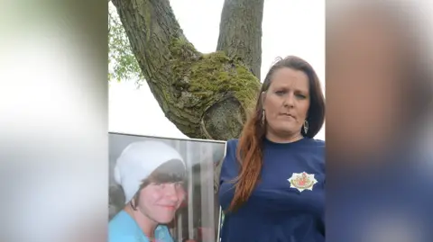 Family photograph Beckie Ramsay with long brown hair wearing a navy blue t-shirt with a water safety logo on it holds a framed photo of her late son, Dylan. He is wearing a white woolly slouch beanie hat and a turquoise t-shirt and smiling. Beckie is standing in front of a tree. She looks impassively. 
