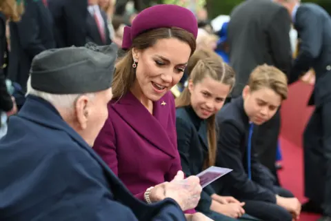 PA Media The Princess of Wales looks at some paper while wearing a plum-coloured dress and hat. She's sitting next to a veteran with her children looking on in the background.