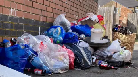 Bags of rubbish piled in a street against a red brick wall.