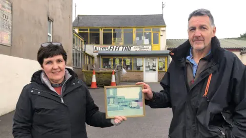 Siblings Susan O'Brien and Melvyn Stroughair holding an old photo of their shop while standing in front of the same shop