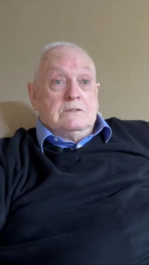 Elderly man with white hair sat in a room wearing blue shirt and jumper