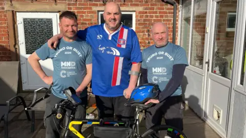 A man with short brown hair with two other men. Two are wearing grey MNDA charity tops and the third is wearing a blue sports top. They all have dark sports trousers. There is a cycle and a cycle helmet and a brick building in the background.