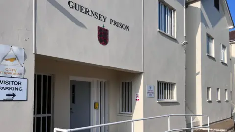 A picture of the front of Guernsey Prison. It is a white building with the logo on the front of it. There is a number of windows and doors
