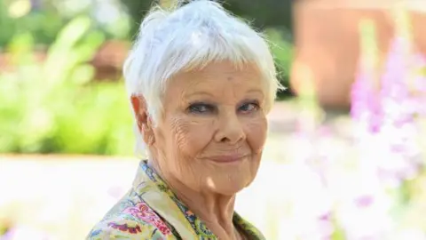 An elderly lady smiles at the camera in a sun-soaked garden