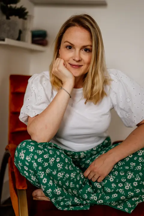 Fables in Focus A woman with blonde, shoulder-length hair. She is sat cross-legged on a chair wearing a white blouse and green floral trousers and is smiling and looking at the camera.