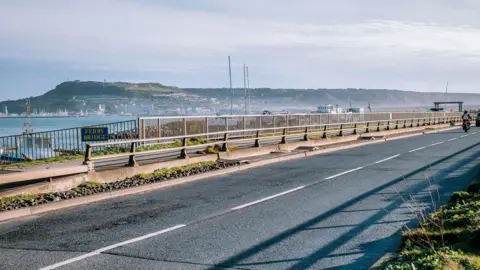 An empty Portland Ferry Bridge - a small bridge crossing a channel at the end of a causeway with railings and a crash barrier separating the footpath from the road.