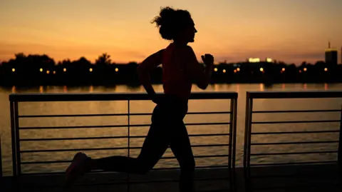The image shows a person jogging along a waterfront promenade during sunset. The silhouette of the runner is clearly visible against the warm orange and pink hues of the sky. In the background, there is a calm body of water reflecting the sunset colours, with a row of trees and distant buildings illuminated by small lights. The foreground features a metal railing that runs parallel to the water, adding structure to the scene.