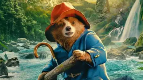 Studiocanal Paddington bear wearing a blue anorak and red hat is holding a tree log which he is presumably using to row in a boat down a fast flowing rainforest river with waterfall in the background. The image is taken from the film Paddington in Peru 