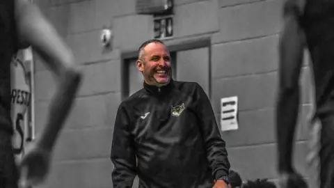 City of Leeds Basketball A man is coaching during a basketball game. He is wearing a black zipped up jacket with an owl logo on, against a brick background.
