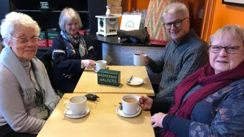 A group of three women and a man are sat around tables at a cafe.  They have cups of coffee in front of them.  There is a green wooden block on the table in front of them which reads 'Wednesday Walkers'.