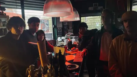 Two men and one woman stand to the left of a table looking at the camera, while three men stand to the right of the table, looking despondent at the camera, in a dark-lit cafe 