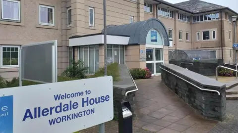 Front entrance of Allerdale House - a two storey building in a light stone colour, with an arched porch with double glass doors in the centre. A paved ramp with low walls on either side leads up to it.
