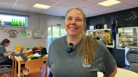 Head and shoulders shot of Faith Rooke-Matthews, wearing a grey t-shirt with blonde hair looking at the camera. The cafe is viable in the background.