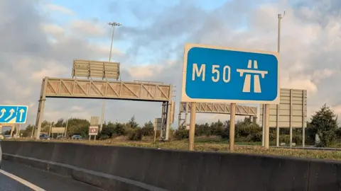 A blue road sign reading M50 on the side of a motorway. 