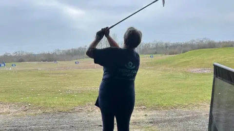 Debbie White An action shot of a woman swinging a golf club on a driving range. The sky is cloudy in the distance. She has the golf club above her head after hitting the ball. The woman is wearing a blue top and leggings. The top has a logo on it that reads "Fight for Sight".