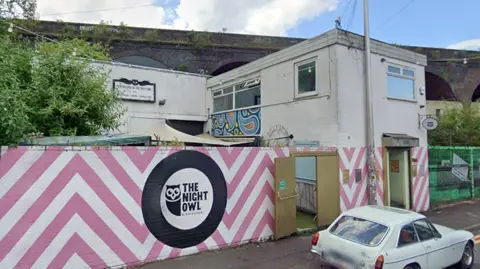 A view of the outside of the Night Owl. A wall at the front is painted white with pink chevrons. The rest of the building is white, with railway arches in the background