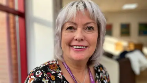 Ann has shoulder-length light hair and is wearing a black, floral dress, with an "NHS eating disorder service" lanyard around her neck. The room behind her is out of focus.